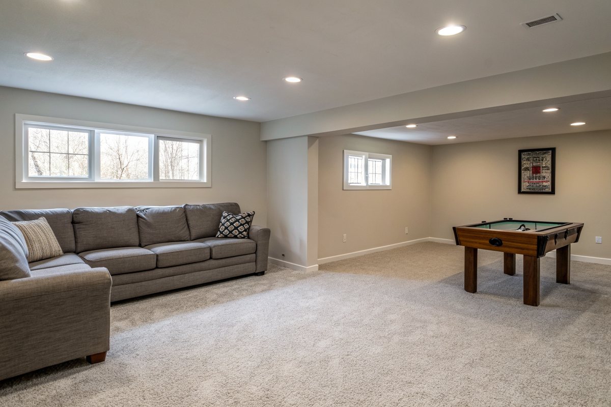 A bright finished basement family room with carpet, a gray sofa and a game table
