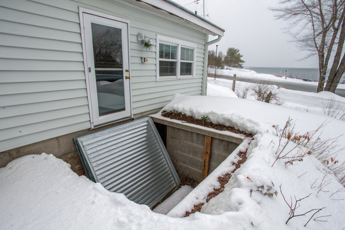 A corrugated metal window well packed with snow beside a clapboard house